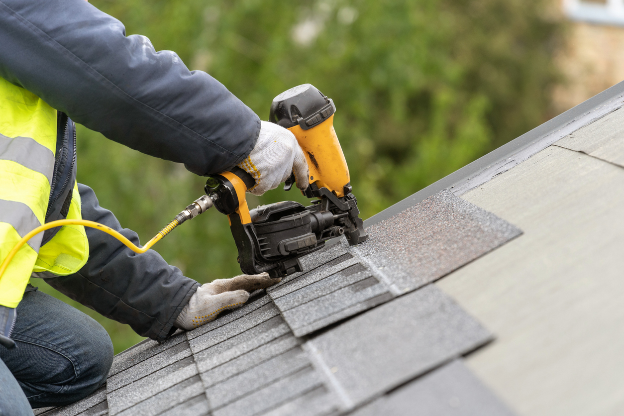 Close-up of a roofer's hands using a pneumatic nail gun to secure shingles on a roof. The worker is wearing a high-visibility jacket with reflective stripes, gloves, and is handling the tool with care. Freshly laid shingles and the blurred greenery of trees in the background suggest an outdoor residential setting during roof installation or repair.